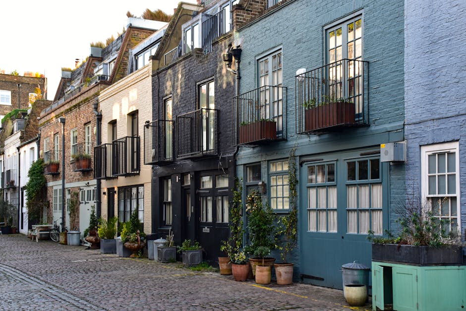 A row of narrow, colorful terraced houses with a mix of pastel blue, black, beige, and white facades along a cobblestone street. Each building features small front garden beds filled with potted plants, shrubs, and climbing greenery, creating a lush, inviting atmosphere. Several houses have small black metal balconies with potted plants, while others have traditional white-framed windows with curtains. The textures of the buildings vary from brick and stucco to painted wood, and the street is lined with potted plants and garden ornaments. Some greenery climbs the walls, adding to the charm of the scene, which is illuminated by soft, natural light, highlighting the textures of the plants, cobblestones, and building facades. The overall scene captures a quaint, well-maintained residential area consistent with garden maintenance and landscaping work, as seen in the context of access challenges at Baker Street mews, with Gardeners Marylebone providing outdoor care services.