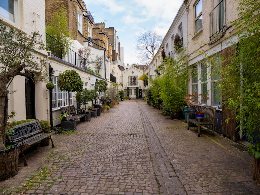A row of narrow, colorful terraced houses with a mix of pastel blue, black, beige, and white facades along a cobblestone street. Each building features small front garden beds filled with potted plants, shrubs, and climbing greenery, creating a lush, inviting atmosphere. Several houses have small black metal balconies with potted plants, while others have traditional white-framed windows with curtains. The textures of the buildings vary from brick and stucco to painted wood, and the street is lined with potted plants and garden ornaments. Some greenery climbs the walls, adding to the charm of the scene, which is illuminated by soft, natural light, highlighting the textures of the plants, cobblestones, and building facades. The overall scene captures a quaint, well-maintained residential area consistent with garden maintenance and landscaping work, as seen in the context of access challenges at Baker Street mews, with Gardeners Marylebone providing outdoor care services.