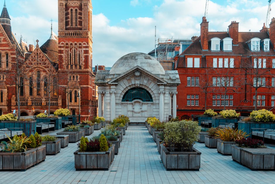 A rooftop garden featuring neatly arranged wooden planters filled with a variety of lush green plants and small flowering shrubs, set on a paved terrace. The garden is bordered by modern blue benches and decorative lighting, with a backdrop of historic red-brick buildings and a church tower. The sky above is partly cloudy with soft sunlight illuminating the scene. The setting illustrates well-maintained garden beds and landscaping work, emphasizing outdoor maintenance and garden care by Gardeners Marylebone, as seen on the page titled 'Best gardeners Marylebone High Street experts near me', hosted at gardenersmarylebone.org.uk.