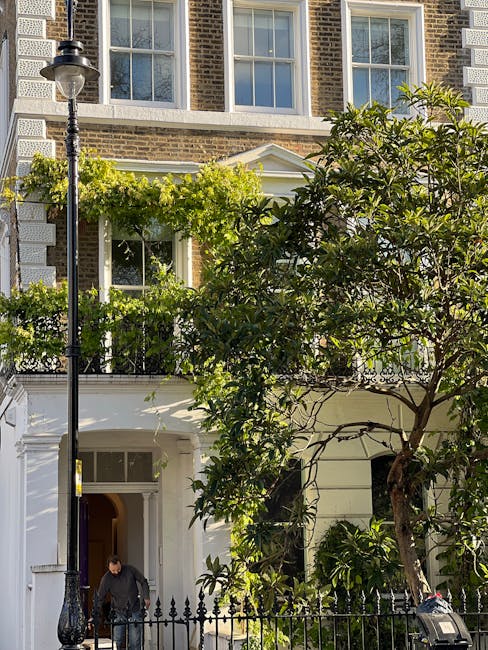 A historic brick townhouse with large sash windows and a white classical façade features a well-maintained garden in front. The garden includes a mature tree with lush green leaves partially covering the building, along with neatly trimmed shrubs and flowering plants along the border. A black metal fence with decorative finials encloses the garden area, and a vintage-style lamp post stands beside the pavement. The lawn appears to be freshly mowed with even, bright green grass, and landscaping tools or maintenance activities, such as pruning or trimming, are not directly visible but are suggested by the clean edges and cultivated appearance. The scene is captured in daytime with natural sunlight illuminating the scene, emphasizing the textures of the brickwork, foliage, and freshly cut grass. This image likely relates to garden maintenance and landscaping work provided by Gardeners Marylebone, demonstrating their expertise in outdoor care near Marylebone High Street.